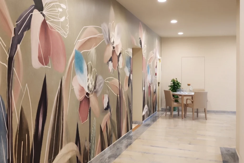 Corridoio di casa di riposo con grande decorazione floreale a parete e, sul fondo, un tavolo con sedute per gli ospiti