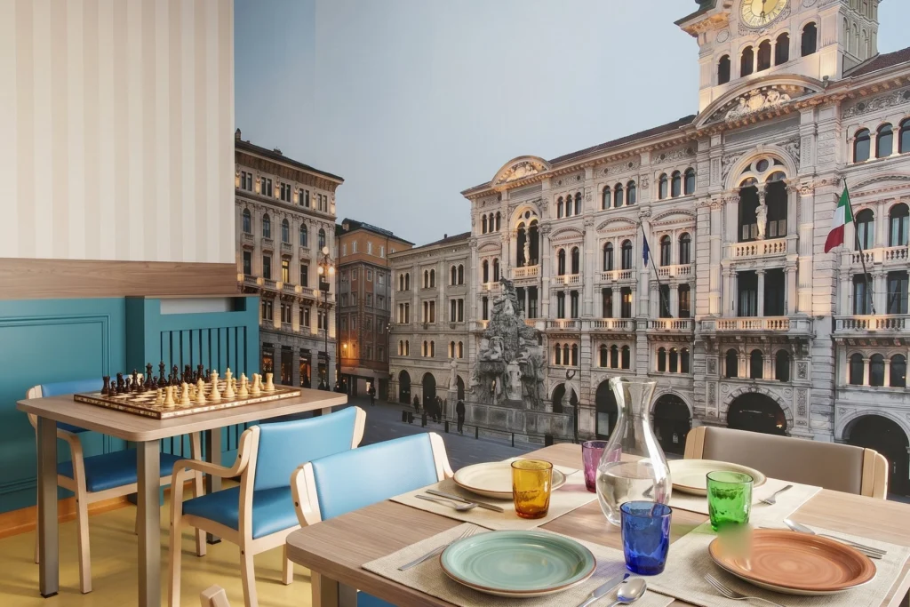 Sala pranzo con murale panoramico, tavoli in legno e sedute imbottite — RSA, progetto Ad Maiores